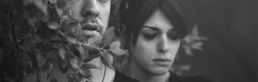 Black and white photo of couple standing in bushes, looking hopelessly into the distance.